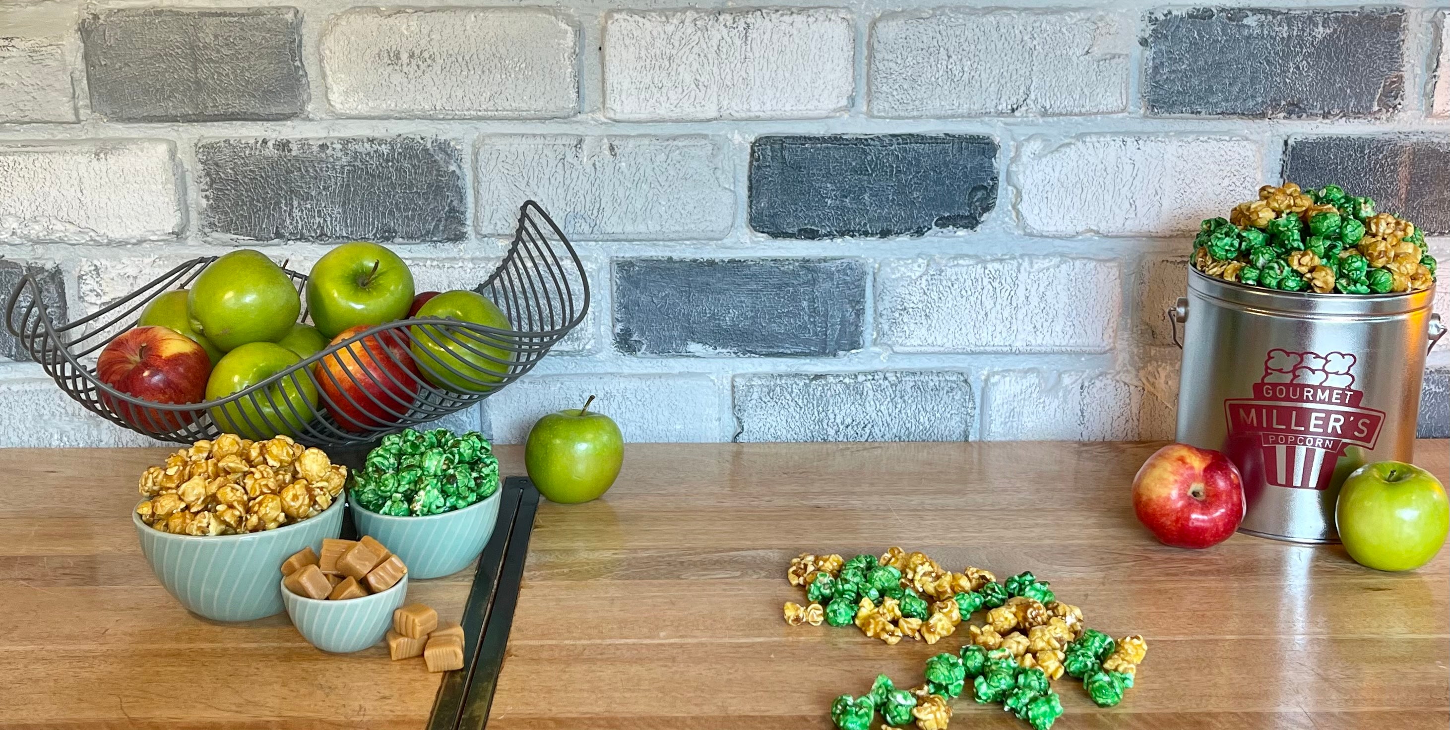 background of grey brick and wood grain counter top. from left to right. fancy wire basket with red and green apples placed behind 3 dishes. 1 with caramel popcorn, 1 with apple popcorn, 1 with caramel squares. blend of the two flavors on the counter top. grey miller's popcorn tin with a blend of the 2 flavors piled above the rim sitting in front of a red apple and a green apple